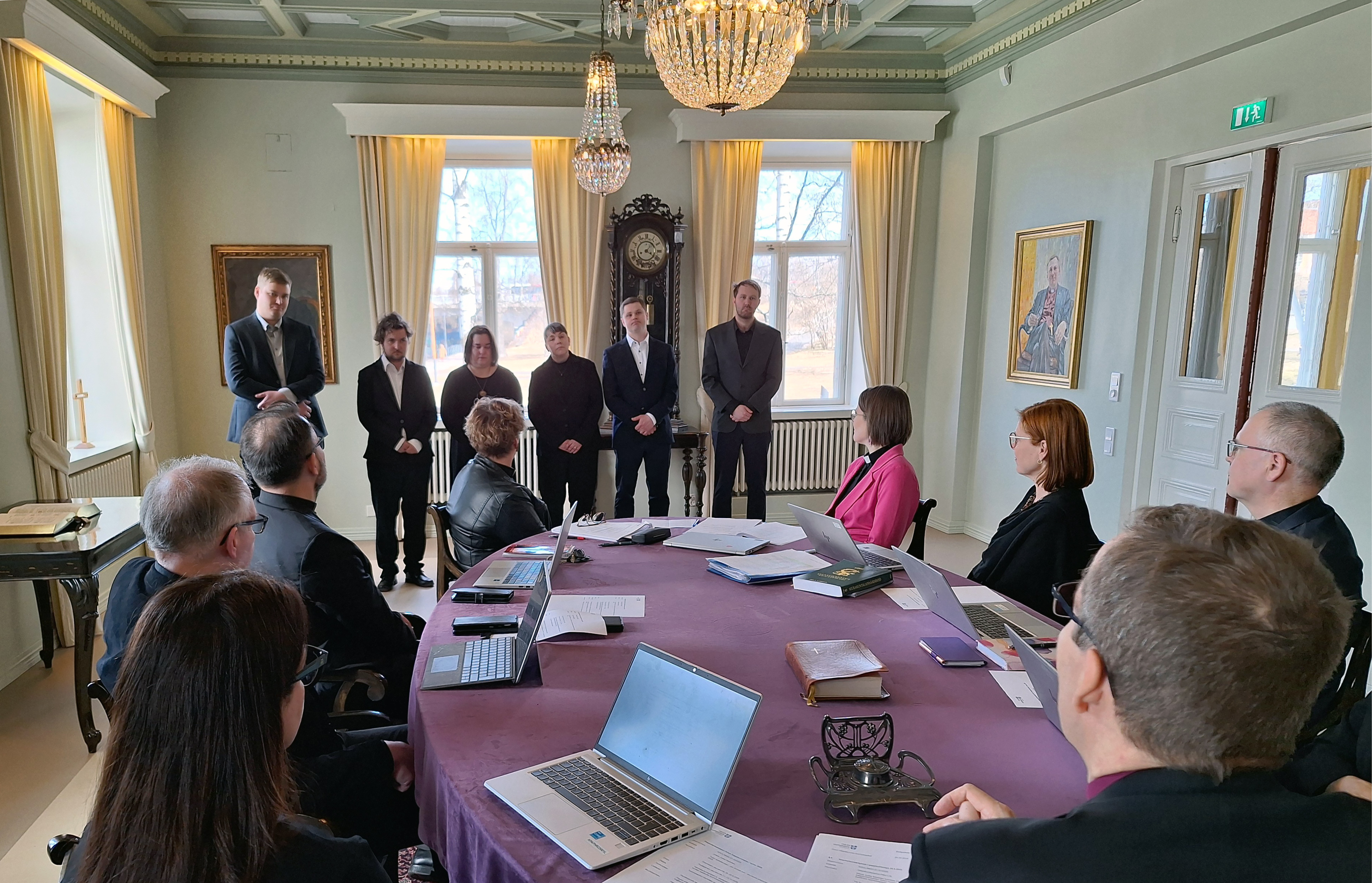 Pöydän ympärillä istuvat ihmiset katsovat rivissä seisovia kuutta henkilöä, jotka vihitään pian papeiksi.