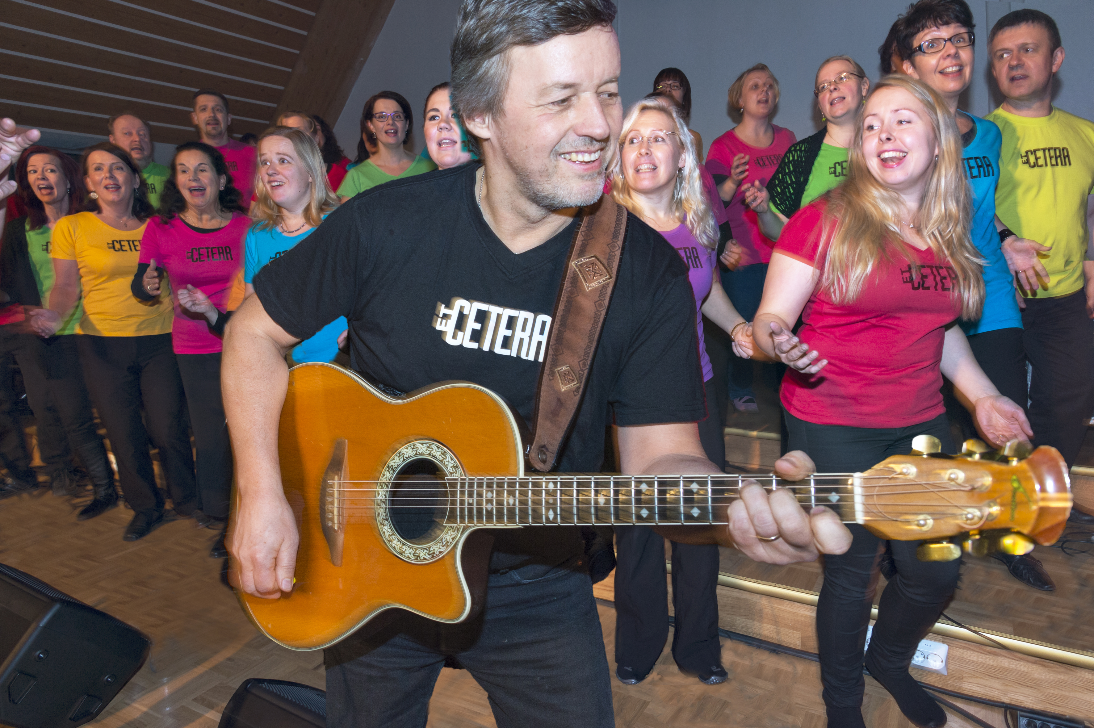 Gospelkuoro EtCetera laulaa innokkaasti taustalla ja edessä kuoron taiteellinen johtaja Pekka Simojoki soittaa hymyillen kitaraa.